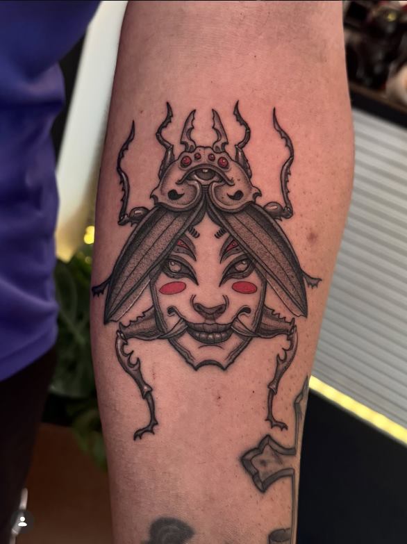Tatuaje fineline de un escarabajo con máscara oni en el antebrazo